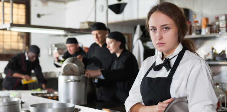 An unhappy waitress stands in a kitchen among other employees.