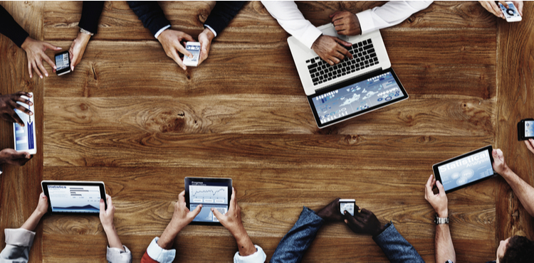 An above view of people sitting around a wooden table on various electronic devices. An above view of people sitting around a wooden table on various electronic devices.