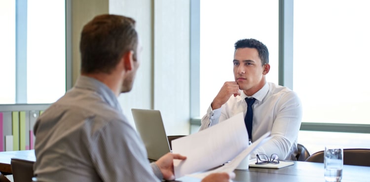 Two men sit on either side of a desk in an office. Two men sit on either side of a desk in an office.