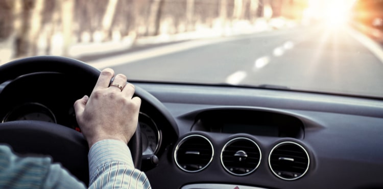 A man's hand on the steering wheel of a car driving down a road.
