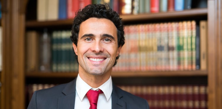 A smiling man in a suit in front of bookshelves. A smiling man in a suit in front of bookshelves.