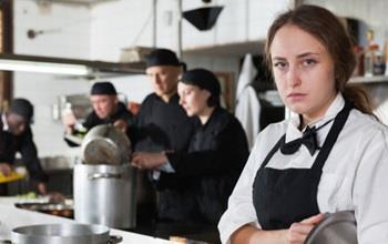 An unhappy waitress stands in a kitchen among other employees.