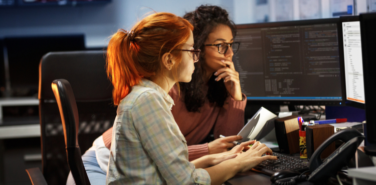 Two women examine code together on a computer display.