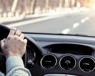 A man's hand on the steering wheel of a car driving down a road.