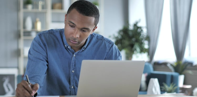 A man writing on a notepad while working on his laptop.