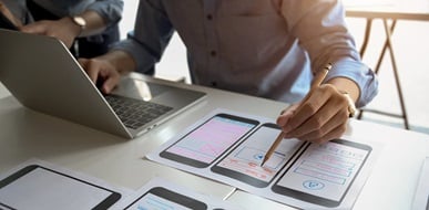 Designer reviewing mobile app on laptop and papers.