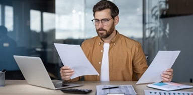Person reviewing documents at desk with laptop in office.