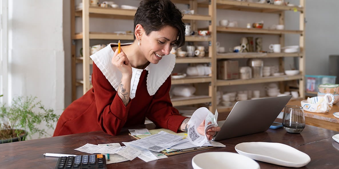 Person reviewing receipts at a table with a laptop in a studio workspace. Person reviewing receipts at a table with a laptop in a studio workspace.