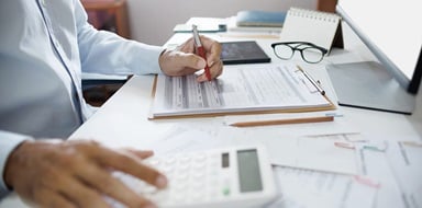 Person filling out tax forms at desk with calculator and laptop.