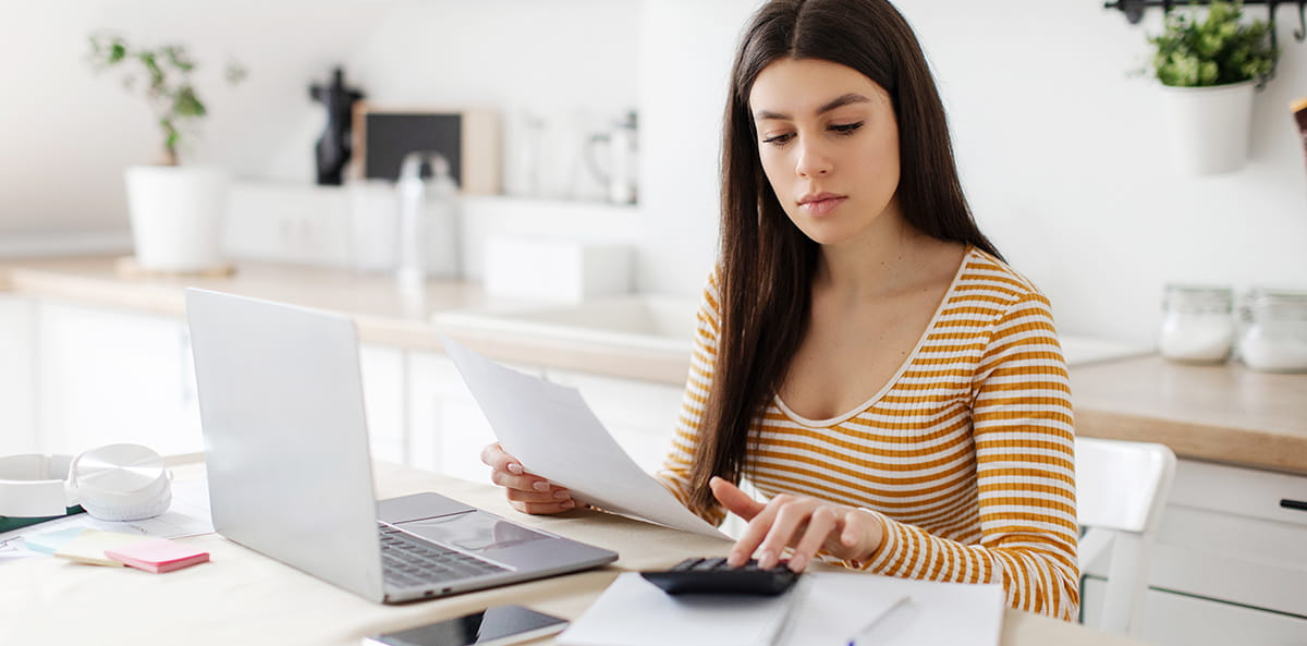 Person working at a kitchen table with a laptop, papers, and a calculator.