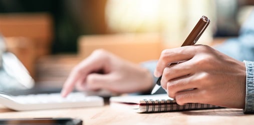 Writing in a notebook while using a calculator at a desk.
