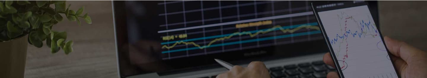 Stockbroker at desk looking at stock charts on a phone and laptop.