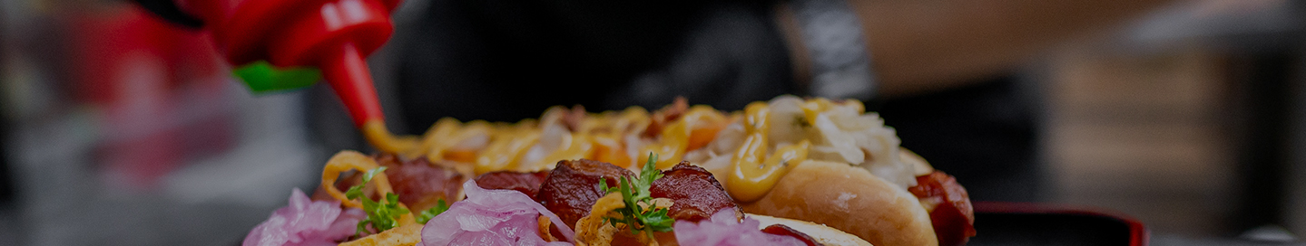 An employee puts condiments on a row of gourmet hot dogs.