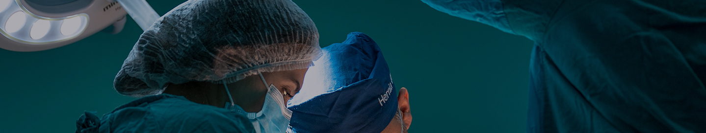 A surgeon in scrubs and a face mask working on a patient.