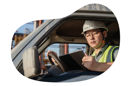 A worker wearing a safety helmet and reflective vest sitting in a vehicle and reviewing a tablet.