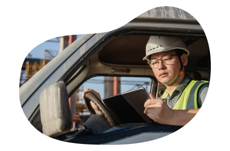 A worker wearing a safety helmet and reflective vest sitting in a vehicle and reviewing a tablet.